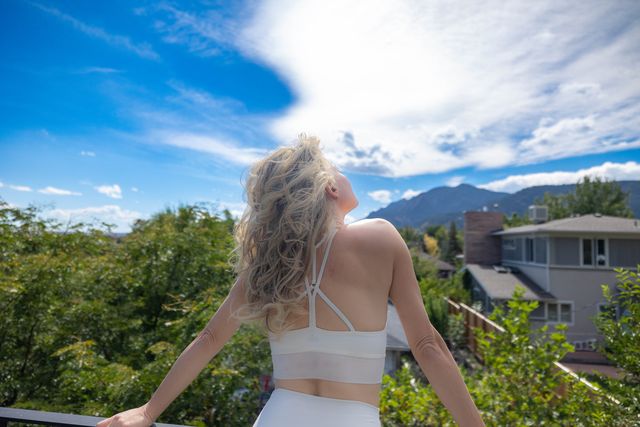 A blond woman is wearing a white two-piece outfit seen from the back, standing with arms outstretched facing a scenic view. The sky is clear with a few clouds, and mountains loom in the distance behind suburban homes. 