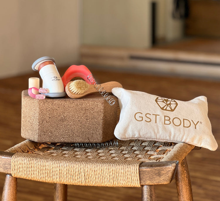 GST Body face-scia kit displayed on a wooden stool. The kit includes a cork block, a dry brush, a lip piece, chin piece, nose piece, and a small canvas clutch labeled ‘GST Body,’ all designed for fascia-focused face exercises.