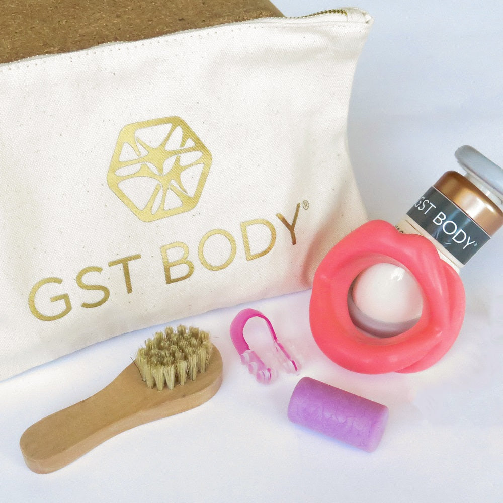 GST Body face-scia kit displayed against a light background, featuring a canvas clutch with the GST Body logo, a wooden dry brush, a pink nose contouring clip, a purple cork decompressor, and a pink lip-shaped resistance tool. The compact clutch holds all the tools for easy portability and storage.