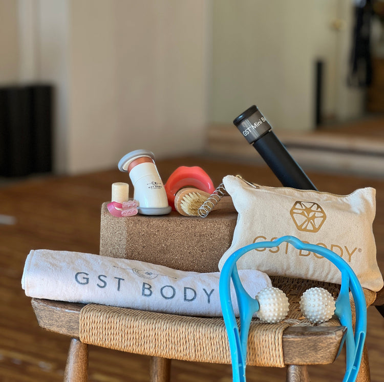 A display of the GST Body Face-scia Kit, featuring a canvas clutch, a GST traction towel, a black mini bar, a cork decompressor, lip and nose tools, a soft bristle brush, and a blue double-ball massage tool. All items are neatly arranged on a stool.