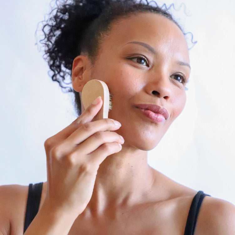 A woman gently using the face dry brush, brushing her cheek in an upward motion. The dry brush stimulates circulation and exfoliates the skin, promoting a healthy, radiant complexion.