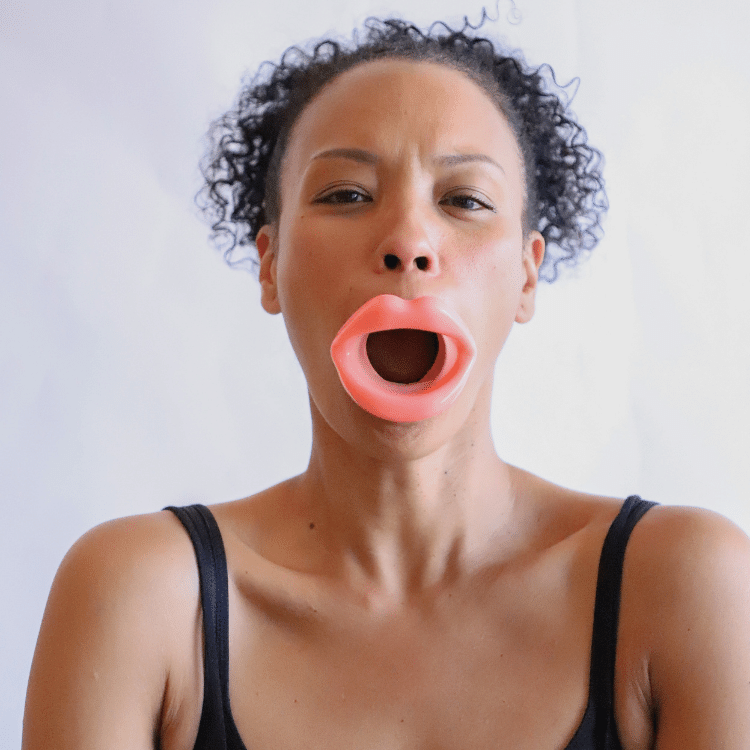 A woman demonstrating the use of the pink Orbital Resistance Band from the GST Body face-scia kit, placed around her mouth. The tool helps exercise facial muscles, particularly around the lips and jaw, promoting muscle engagement and toning.